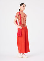 A woman stands against a plain white background, wearing a peach and red patterned top, red wide-leg pants, white shoes, and carrying the LEAF PLEATS BAG by me ISSEY MIYAKE.