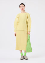 A woman with straight dark hair stands against a white background, wearing a light yellow ribbed sweater and skirt, green socks, beige shoes, and holding the LOGO PLEATS BAG by me ISSEY MIYAKE.