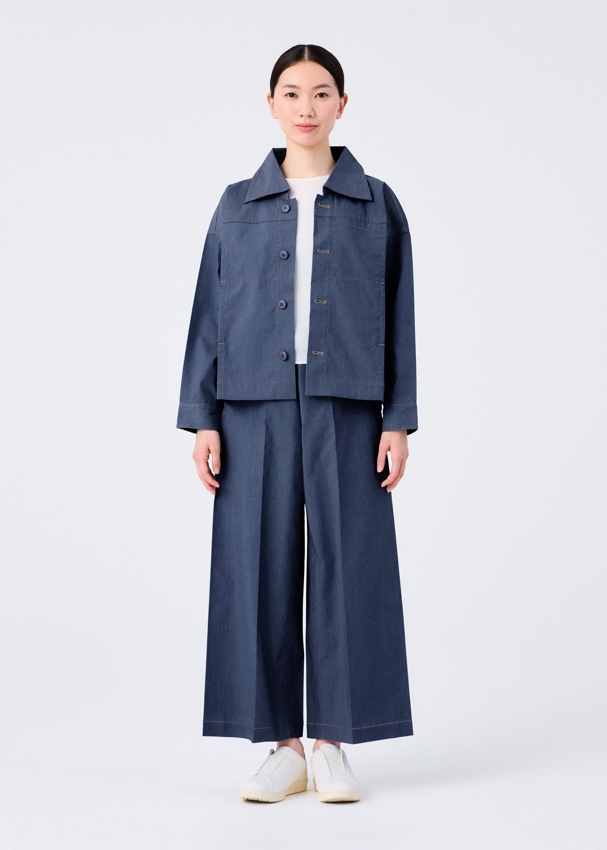 A person faces forward in a loose-fitting FLAT DENIM jacket and matching wide-leg pants from me ISSEY MIYAKE, featuring a recycled polyester denim look, styled with a white shirt and slip-on shoes against a plain white background. view 6 of 7