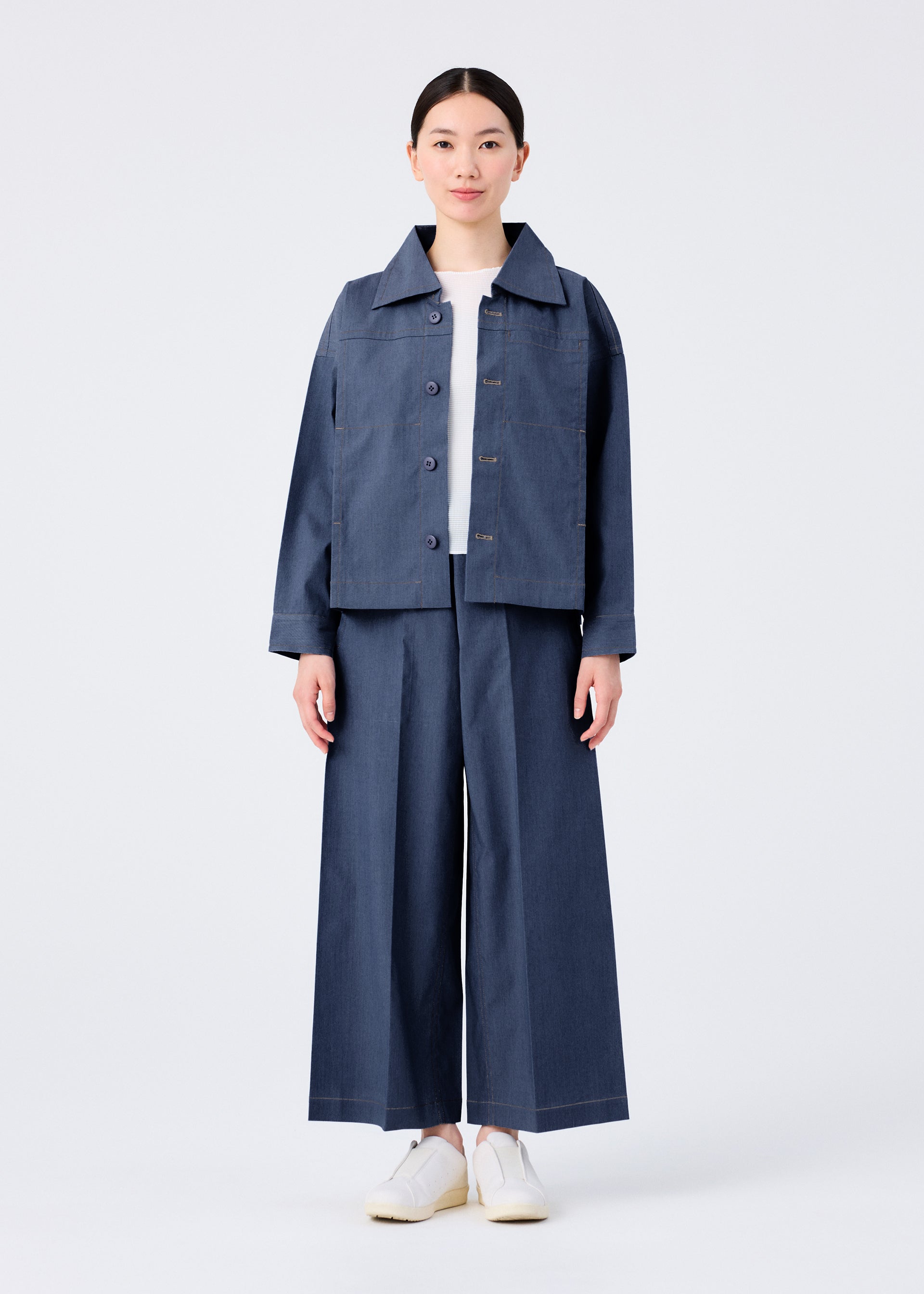 Zoomed view A person faces forward in a loose-fitting FLAT DENIM jacket and matching wide-leg pants from me ISSEY MIYAKE, featuring a recycled polyester denim look, styled with a white shirt and slip-on shoes against a plain white background.