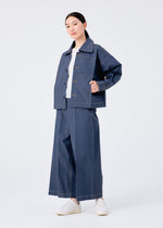 A person poses confidently against a white background wearing the FLAT DENIM portable jacket and wide-leg pants by me ISSEY MIYAKE, crafted from recycled polyester denim, paired with white shoes and one hand in their pocket.