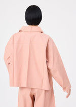 A person with short black hair, seen from behind, wears a loose-fitting FLAT DENIM SEASONAL jacket and matching pants in salmon pink recycled polyester denim by me ISSEY MIYAKE against a plain white background.