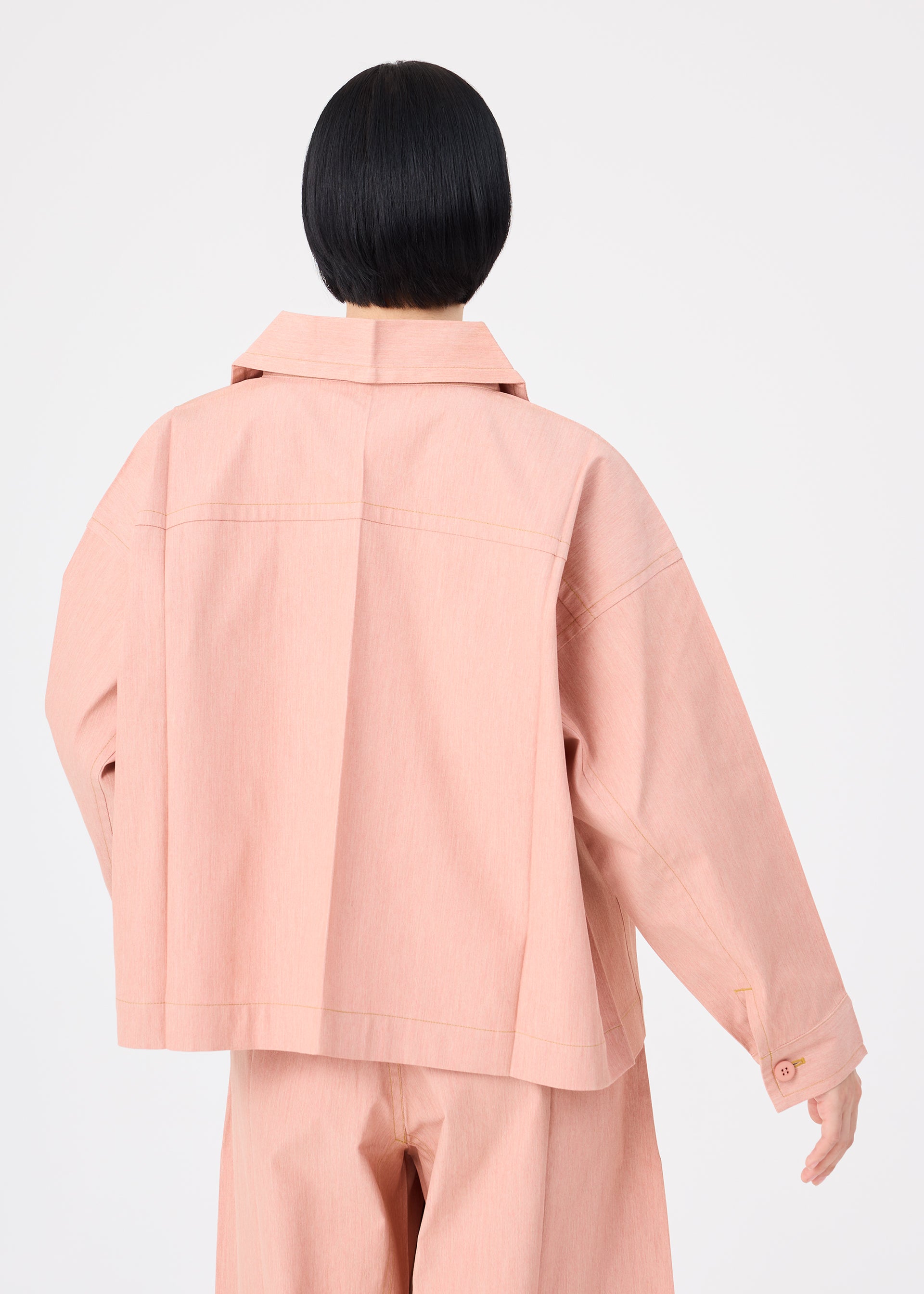 Zoomed view A person with short black hair, seen from behind, wears a loose-fitting FLAT DENIM SEASONAL jacket and matching pants in salmon pink recycled polyester denim by me ISSEY MIYAKE against a plain white background.