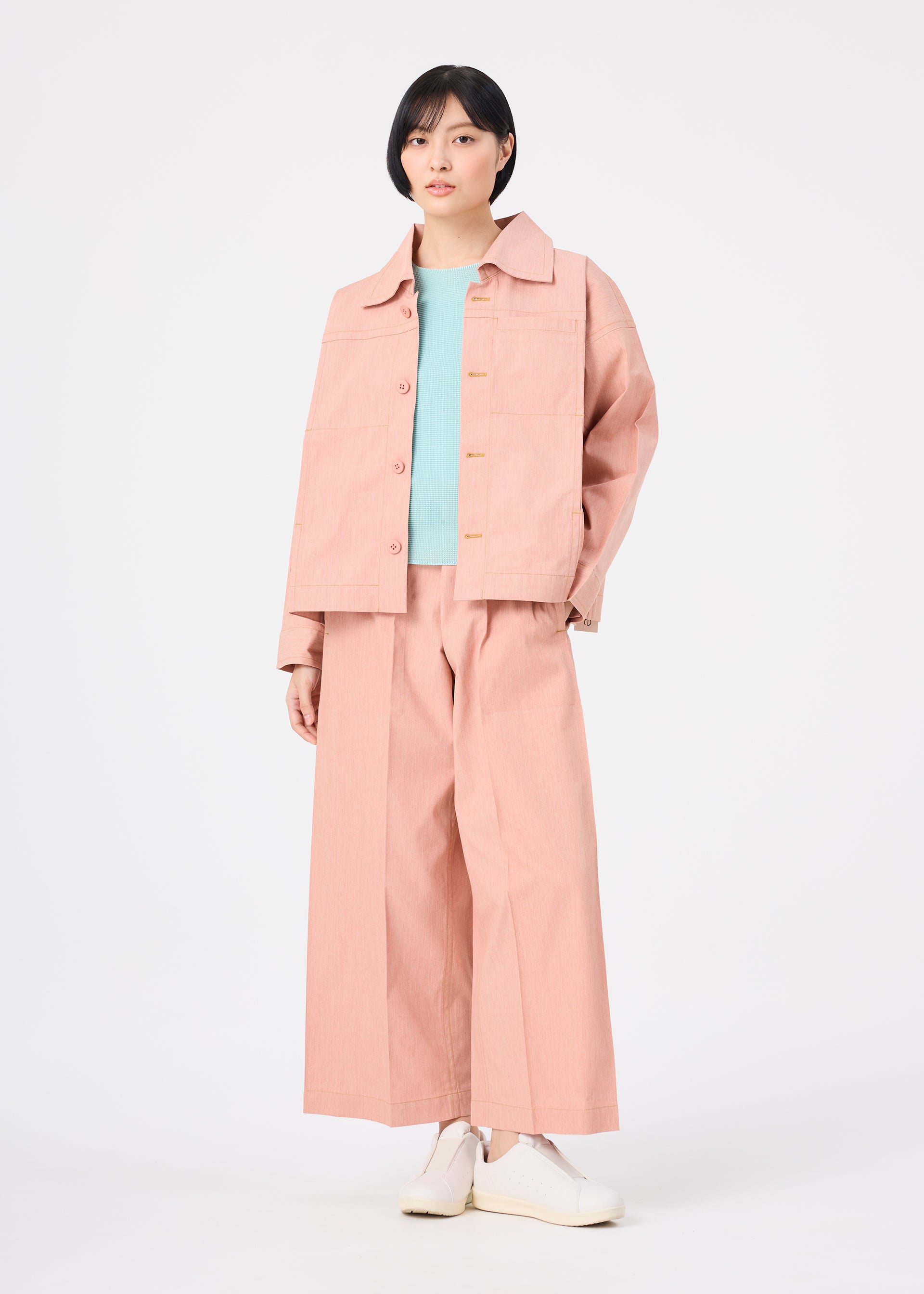 Zoomed view A person with short black hair models the FLAT DENIM SEASONAL wide-legged pants by me ISSEY MIYAKE, styled with a salmon pink portable jacket, light blue top, and white shoes against a plain white background.