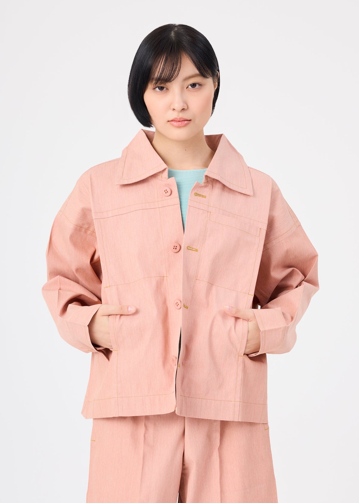 A person with short black hair wears me ISSEY MIYAKE's FLAT DENIM SEASONAL salmon pink portable jacket and matching pants, made from recycled polyester denim, standing with hands in pockets against a plain white background. view 7 of 7