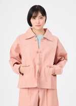 A person with short black hair wears me ISSEY MIYAKE's FLAT DENIM SEASONAL salmon pink portable jacket and matching pants, made from recycled polyester denim, standing with hands in pockets against a plain white background.