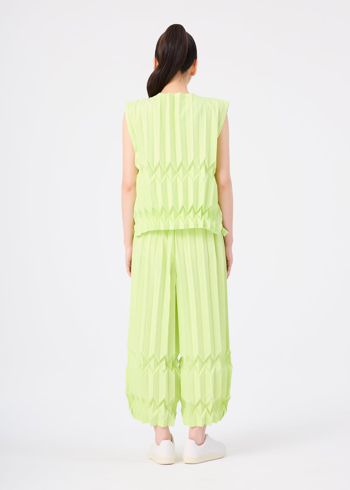 A person faces away, wearing the TREE PLEATS set by me ISSEY MIYAKE—a light green, sleeveless top and matching wide pants with hand pleating—styled with a ponytail and white slip-on shoes against a plain white background. view 6 of 6