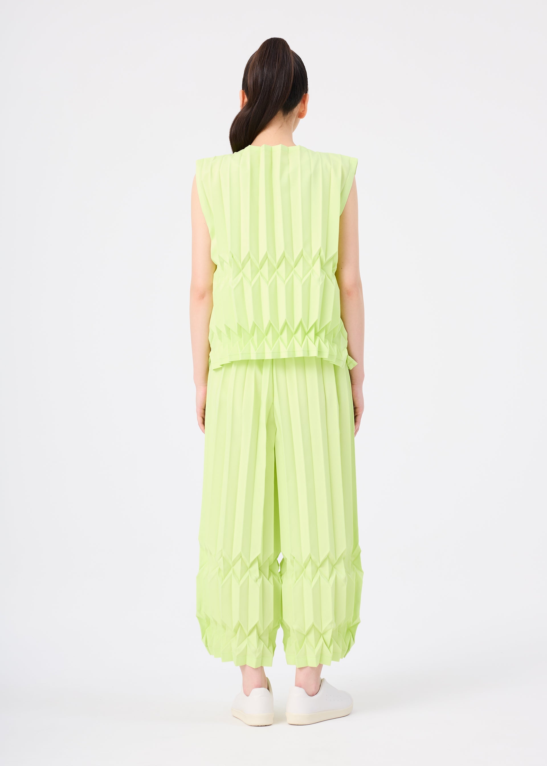 Zoomed view A person faces away, wearing the TREE PLEATS set by me ISSEY MIYAKE—a light green, sleeveless top and matching wide pants with hand pleating—styled with a ponytail and white slip-on shoes against a plain white background.