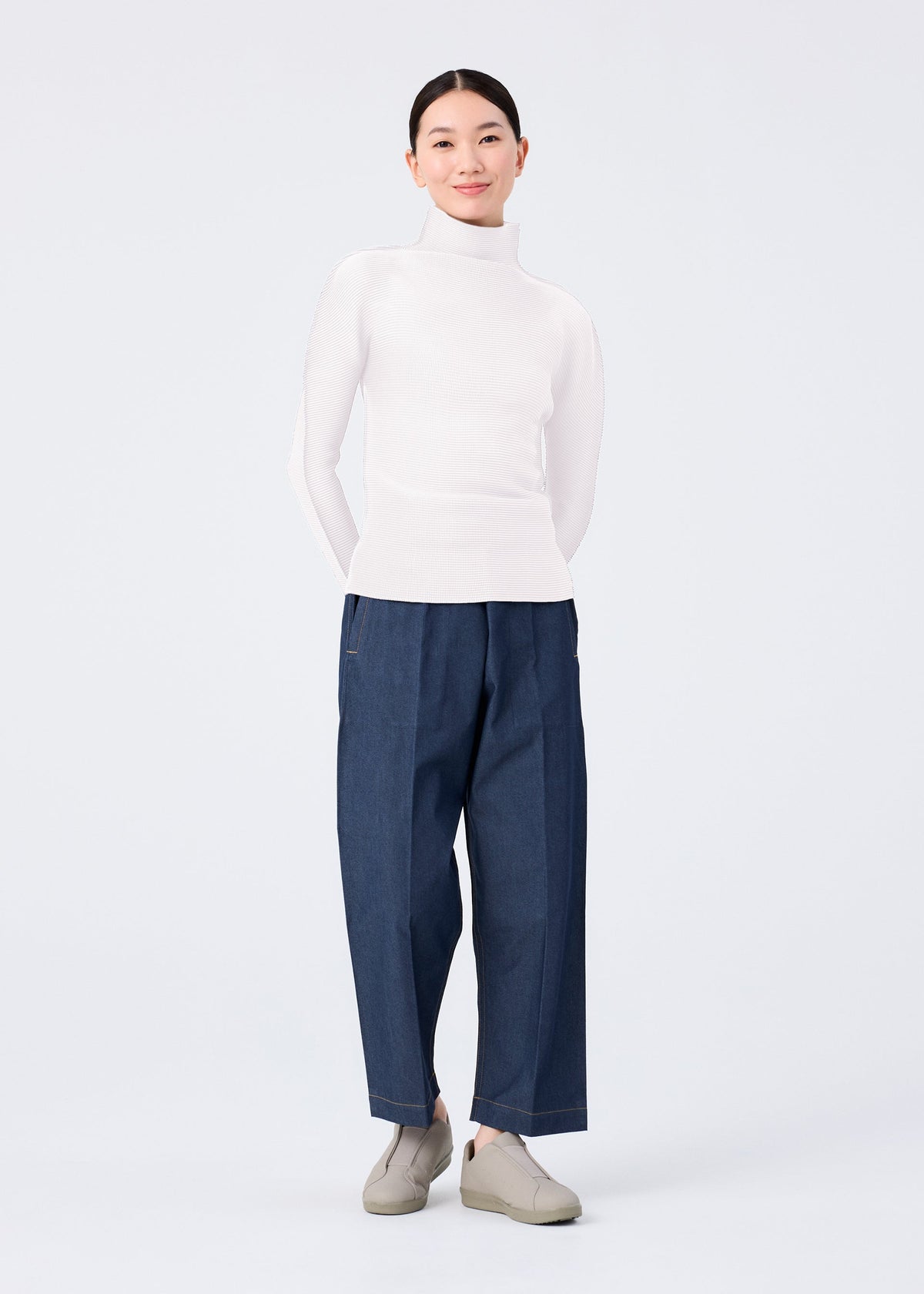 A person stands against a plain background, hands in pockets and smiling slightly, wearing me ISSEY MIYAKE FLAT DENIM loose straight pants, a white turtleneck sweater, and light gray slip-on shoes. view 4 of 5