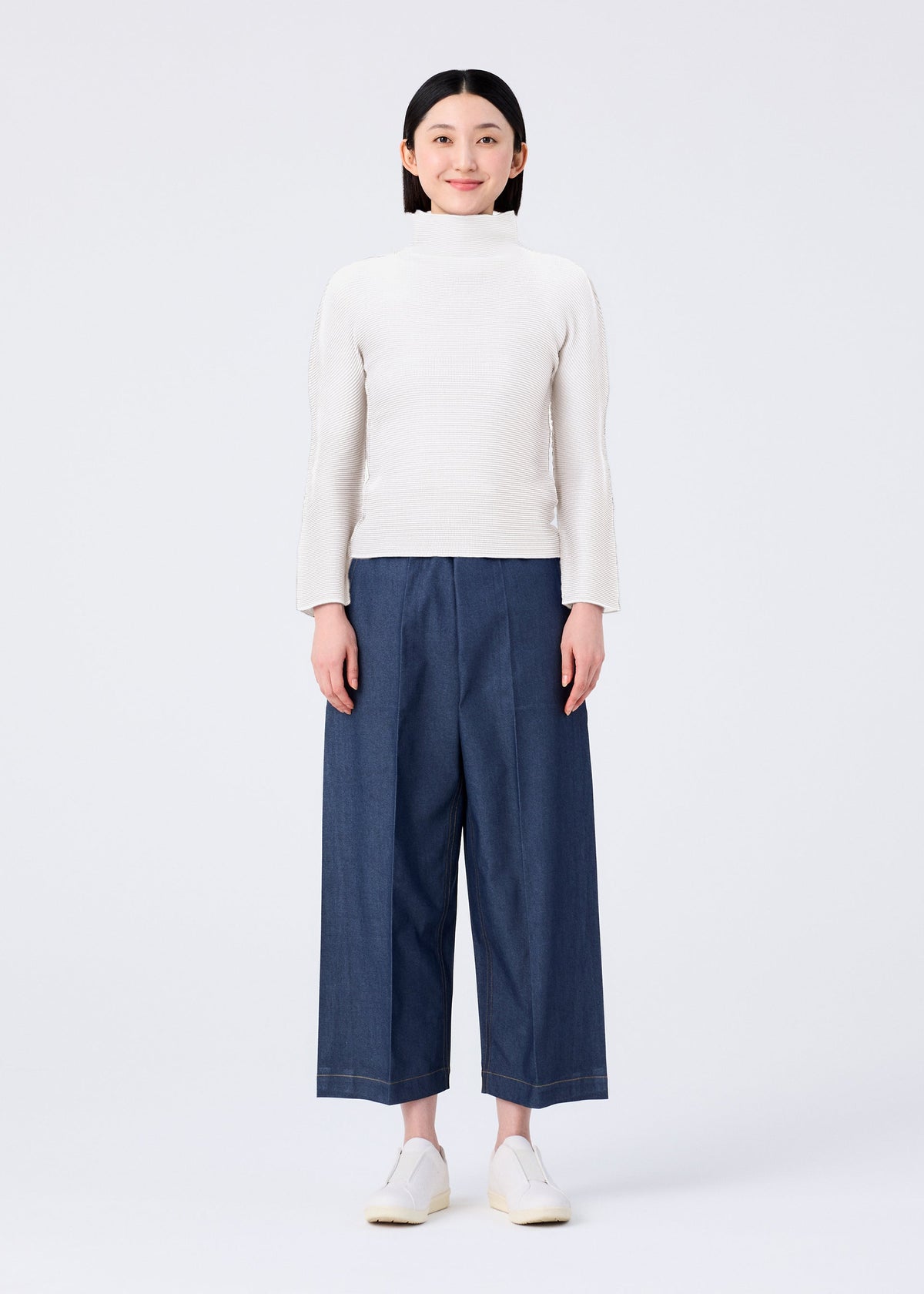 A person stands facing forward, smiling, dressed in a white long-sleeve top, FLAT DENIM wide pants by me ISSEY MIYAKE, and white slip-on shoes against a plain white background. view 4 of 5