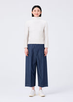 A person stands facing forward, smiling, dressed in a white long-sleeve top, FLAT DENIM wide pants by me ISSEY MIYAKE, and white slip-on shoes against a plain white background.