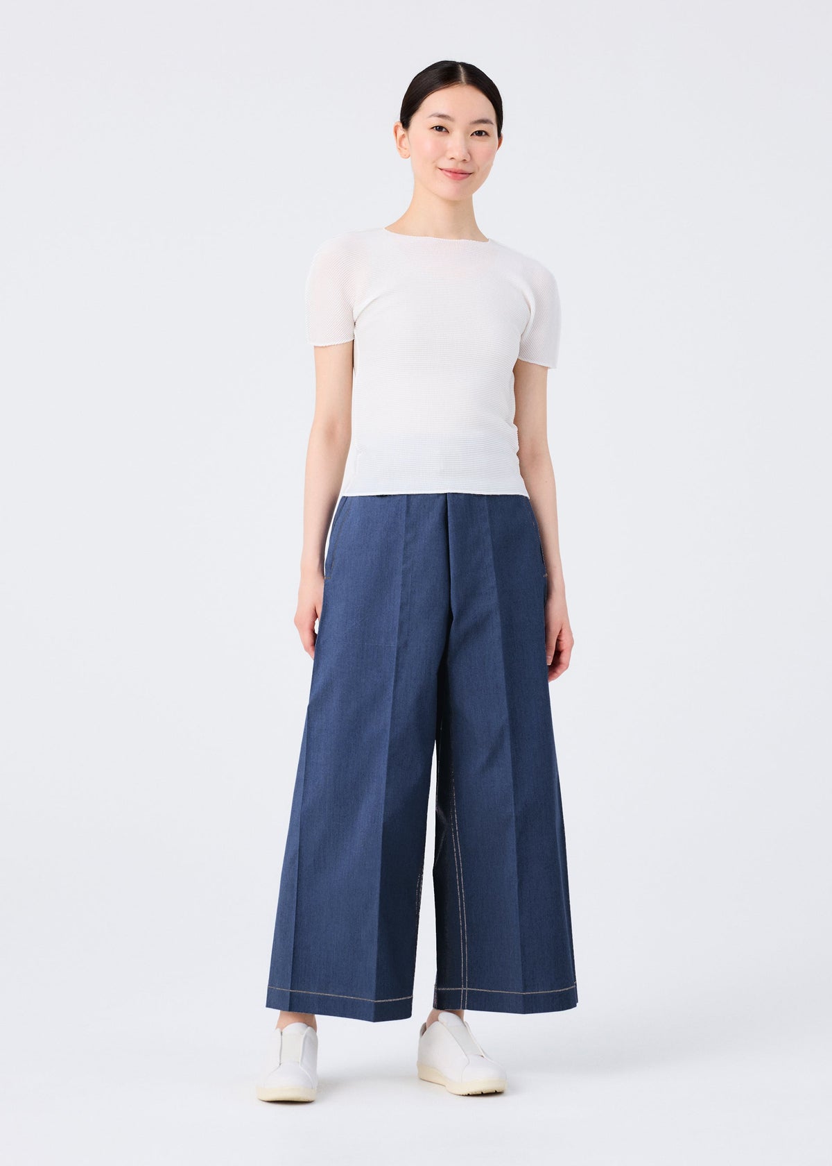 A person stands against a plain white background, wearing a short-sleeved white top, FLAT DENIM wide pants by me ISSEY MIYAKE made from recycled polyester denim with a realistic texture, and white slip-on shoes. Their hair is pulled back and they smile softly. view 4 of 5