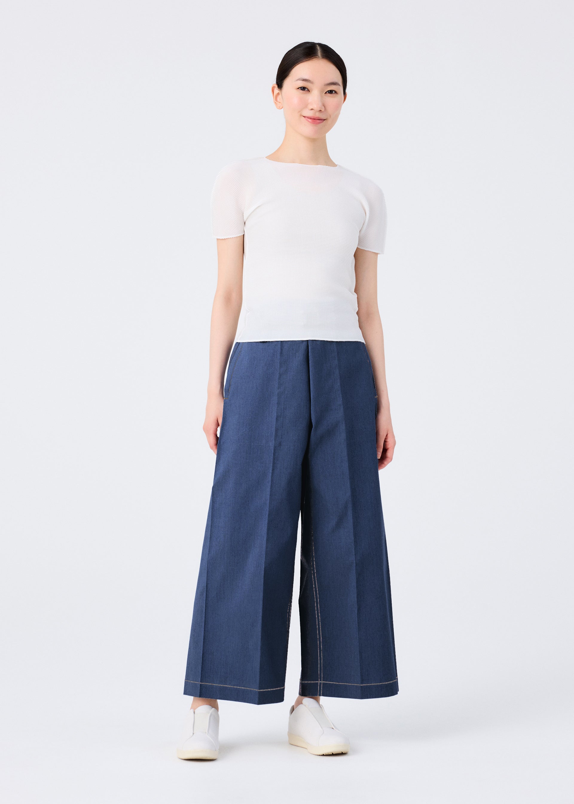 Zoomed view A person stands against a plain white background, wearing a short-sleeved white top, FLAT DENIM wide pants by me ISSEY MIYAKE made from recycled polyester denim with a realistic texture, and white slip-on shoes. Their hair is pulled back and they smile softly.