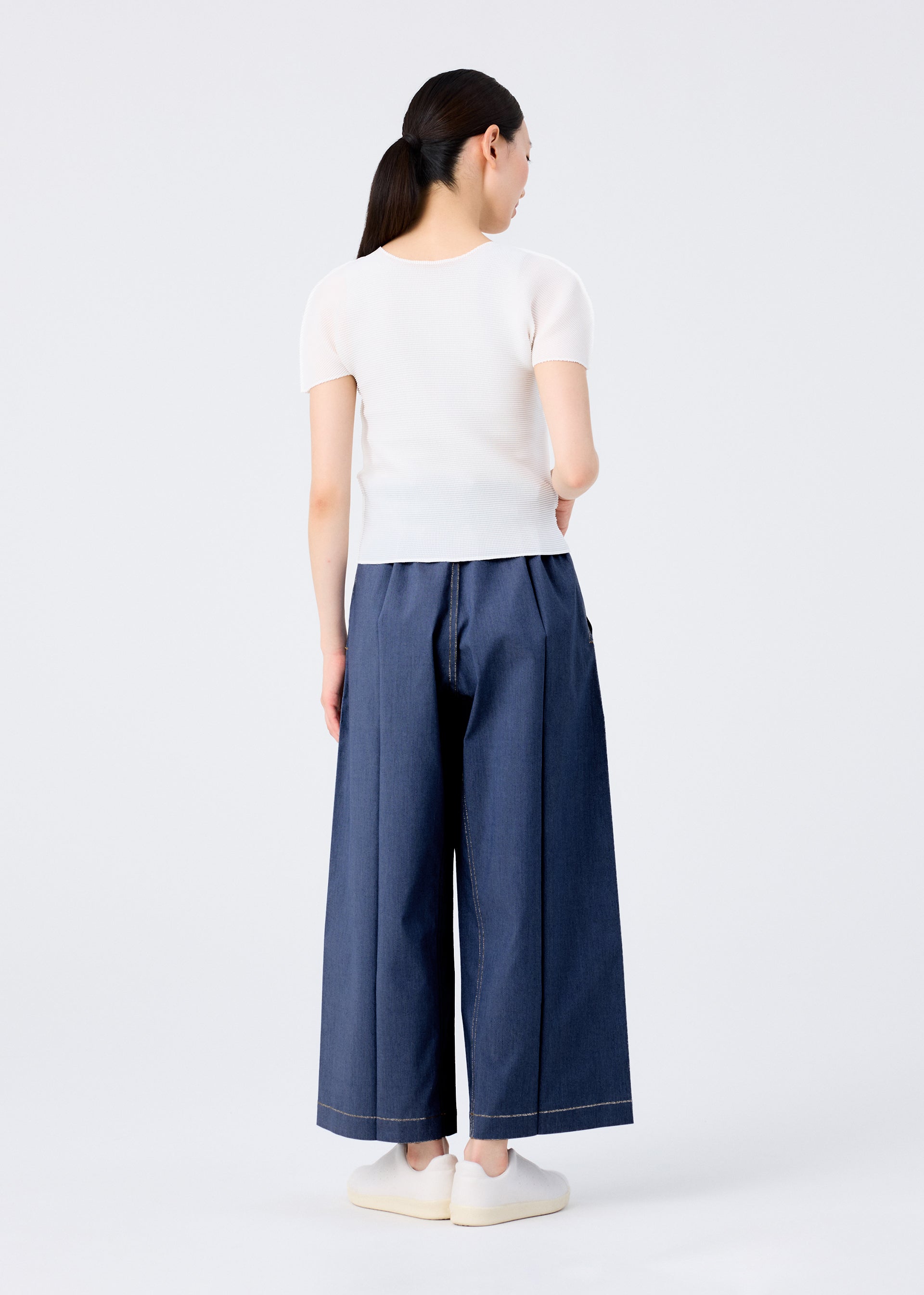 Zoomed view A woman with straight dark hair in a ponytail, wearing FLAT DENIM wide-leg blue pants by me ISSEY MIYAKE, a short-sleeved white top, and white slip-on shoes, stands facing away against a plain white background.