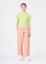 A person stands facing forward against a plain white background, wearing me ISSEY MIYAKE’s FLAT DENIM SEASONAL salmon pink straight pants in recycled polyester and a light green short-sleeve top with white slip-on shoes.