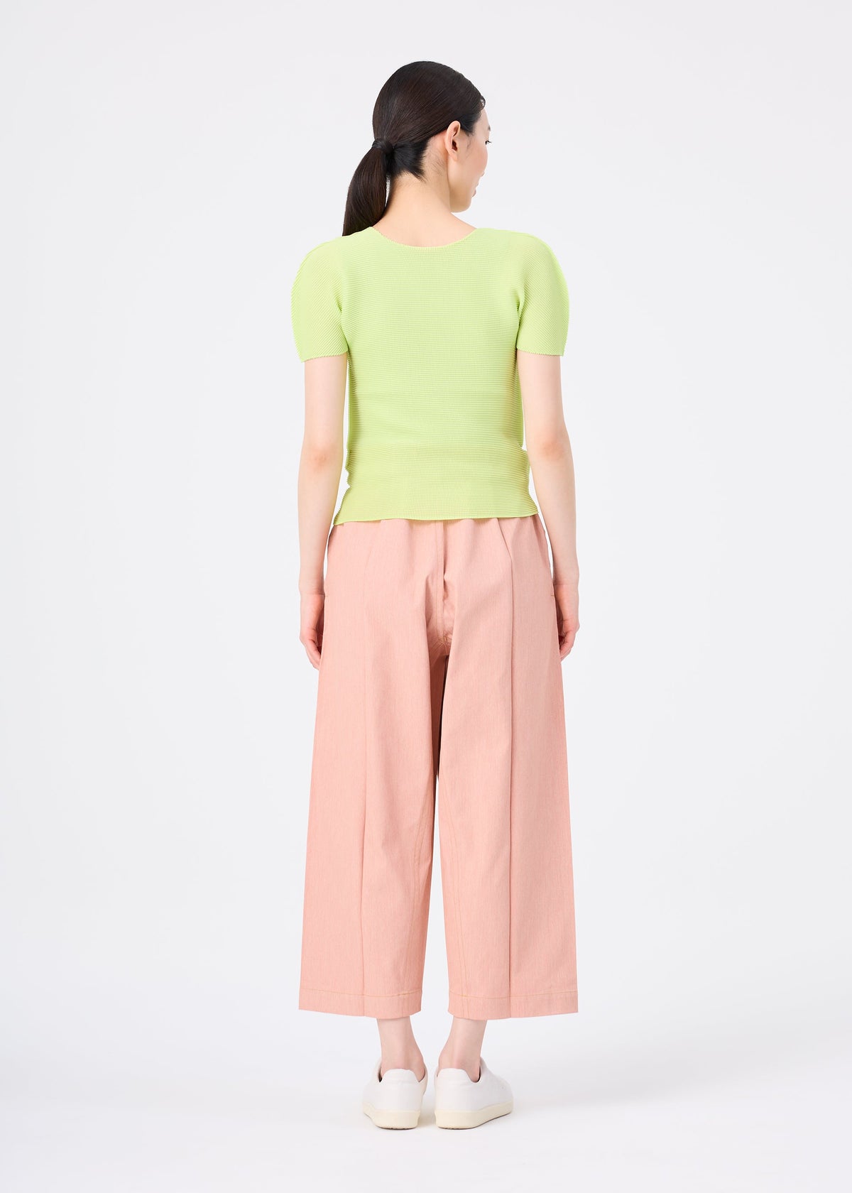 A woman, seen from behind against a white background, wears me ISSEY MIYAKE's FLAT DENIM SEASONAL wide-legged salmon pink pants with a light green ribbed top and white slip-on shoes. view 5 of 5