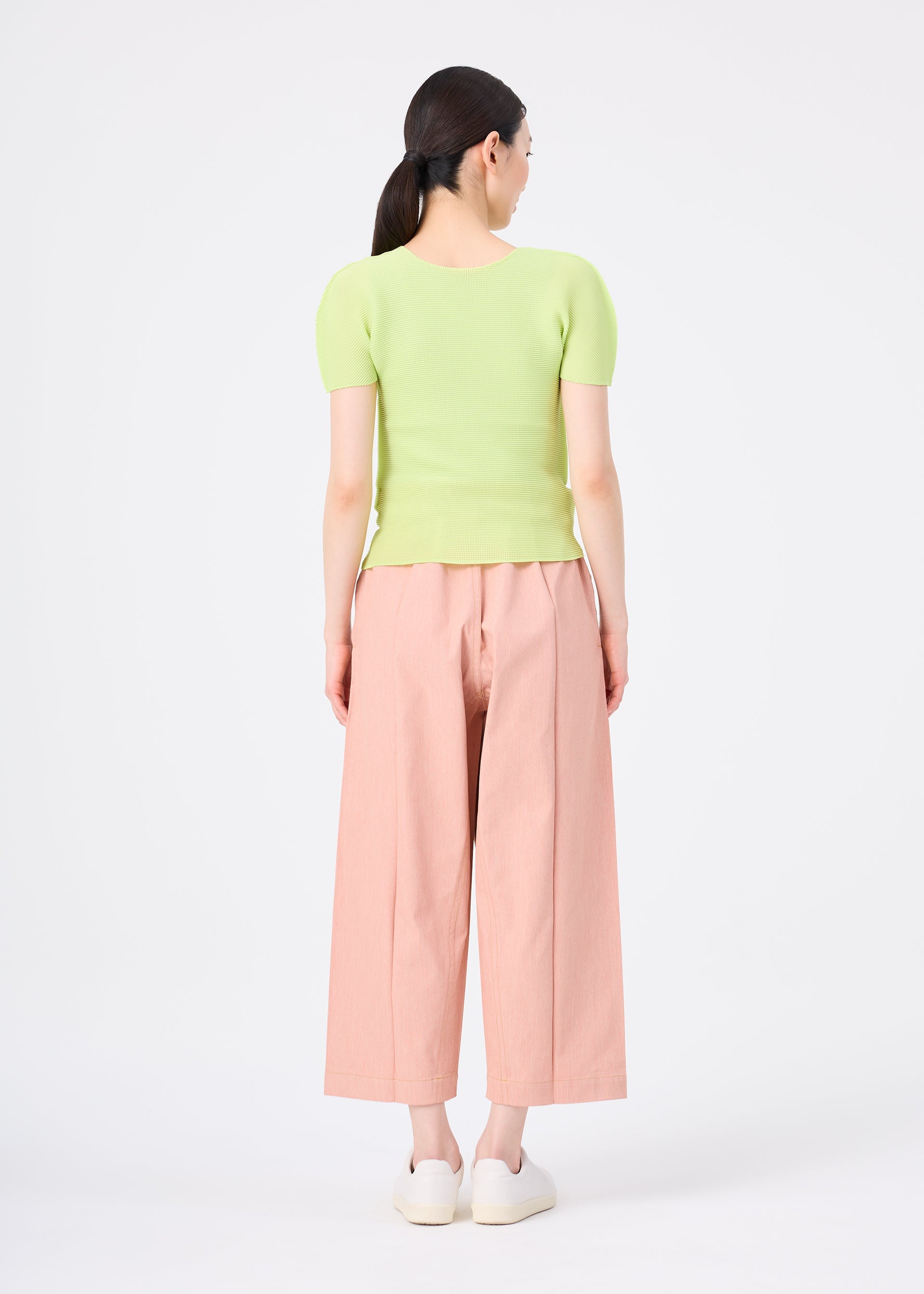Zoomed view A woman, seen from behind against a white background, wears me ISSEY MIYAKE's FLAT DENIM SEASONAL wide-legged salmon pink pants with a light green ribbed top and white slip-on shoes.