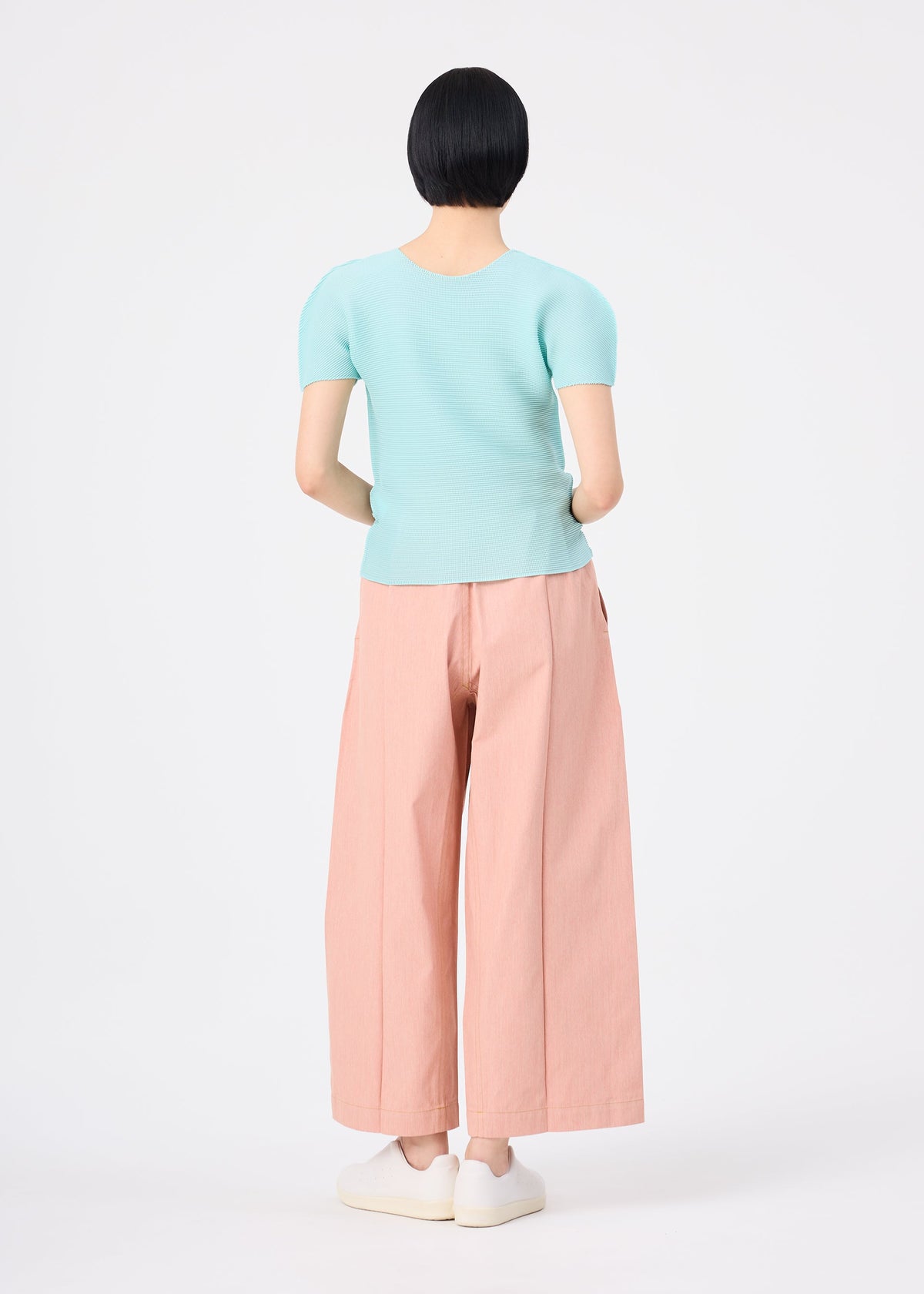 A person with short black hair stands facing away, wearing a light blue short-sleeve top, white shoes, and salmon pink FLAT DENIM SEASONAL pants by me ISSEY MIYAKE against a plain white background. view 5 of 5