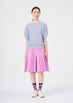 A woman stands facing forward against a plain white background, wearing a pale blue top, the TOWER PLEATS BOTTOM by me ISSEY MIYAKE—handmade pleated shorts in a soft pink hue that mimic the look of a skirt—patterned purple socks, and white shoes.