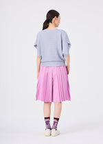 A woman stands facing away, wearing a light blue short-sleeve sweater, the me ISSEY MIYAKE TOWER PLEATS BOTTOM in soft pink featuring an accordion-inspired pleated design, purple patterned socks, and white shoes against a plain white background.