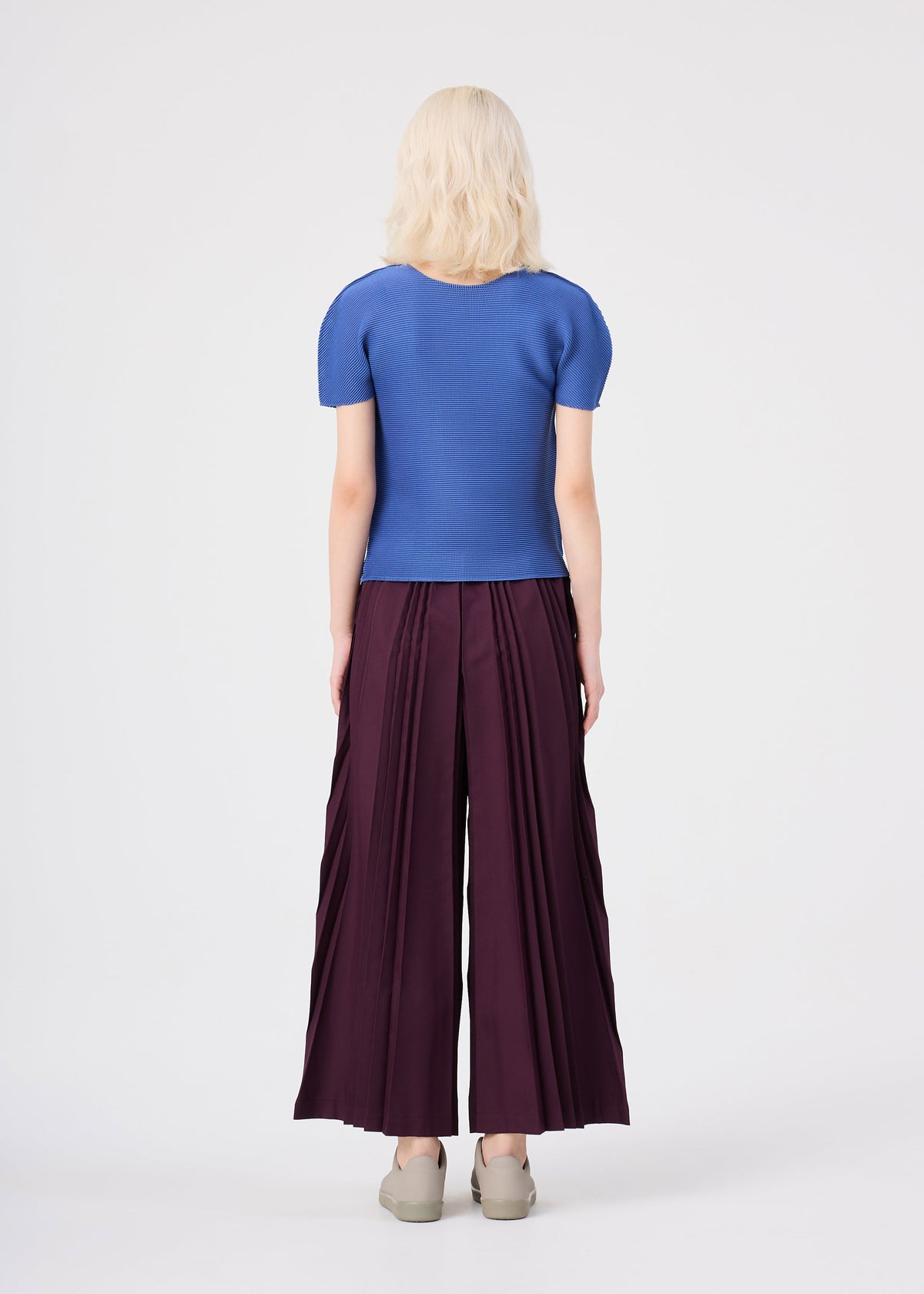 A person with light hair stands facing away, dressed in a blue short-sleeved top and the TOWER PLEATS BOTTOM by me ISSEY MIYAKE—wide maroon pants featuring handmade pleating—paired with beige shoes, against a plain white background. view 7 of 7