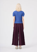 A person with light hair stands facing away, dressed in a blue short-sleeved top and the TOWER PLEATS BOTTOM by me ISSEY MIYAKE—wide maroon pants featuring handmade pleating—paired with beige shoes, against a plain white background.