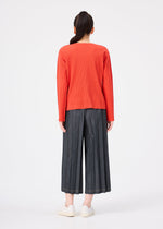A person stands facing away, wearing a vibrant orange long-sleeve top paired with the DENIM PLEATS BOTTOM by me ISSEY MIYAKE—wide-leg, hand-pleated gray pants crafted from recycled polyester—and crisp white sneakers.