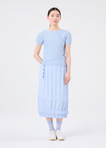 A woman faces forward against a white background, wearing the TREE PLEATS short-sleeve top and matching hand-pleated geometric skirt by me ISSEY MIYAKE, paired with blue socks and white shoes.