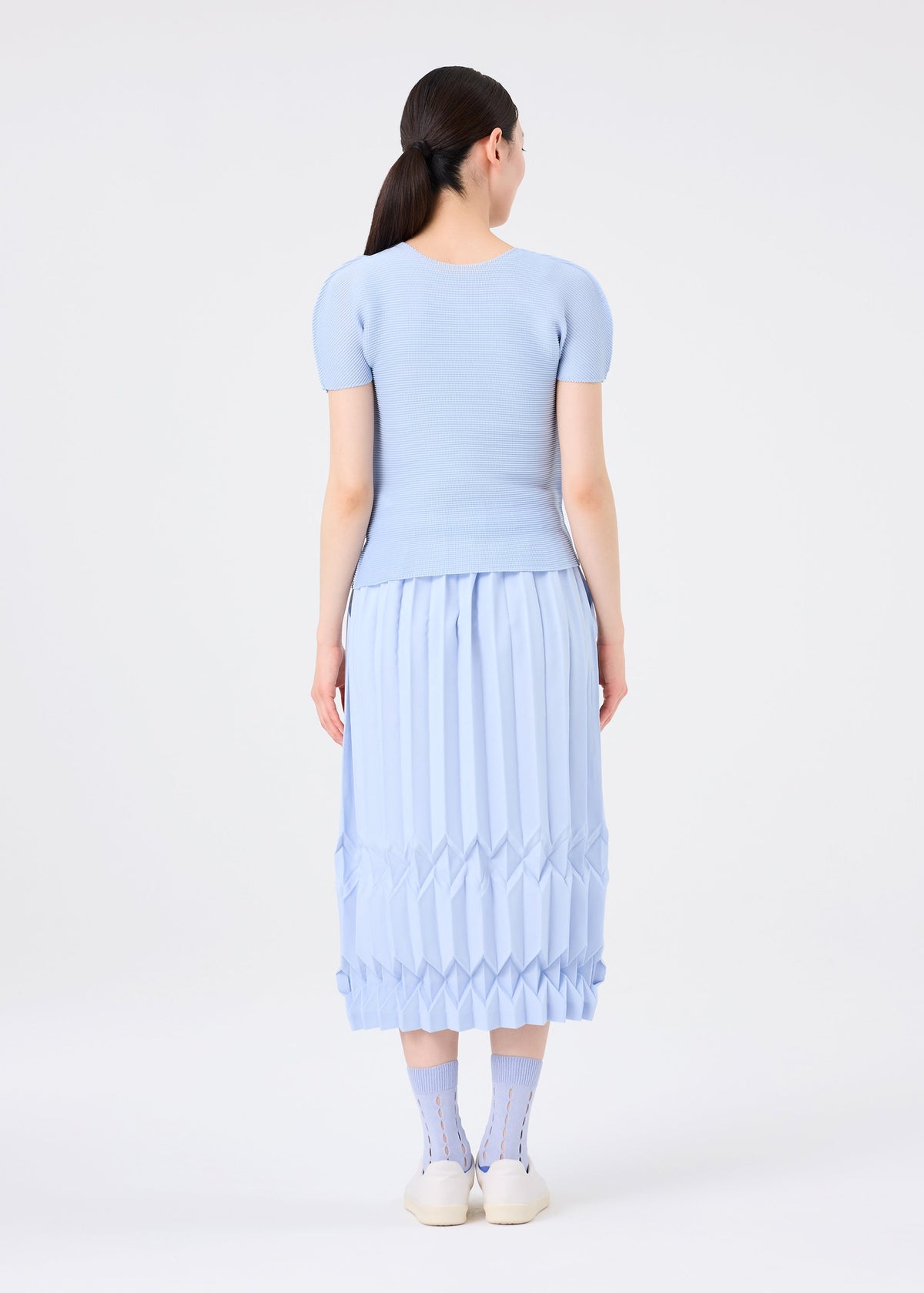 A woman with long dark hair in a low ponytail stands facing away, wearing the TREE PLEATS hand-pleated light blue midi skirt by me ISSEY MIYAKE, a matching short-sleeved top, patterned socks, and white shoes against a plain white background. view 7 of 8