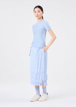 A woman wears the TREE PLEATS skirt by me ISSEY MIYAKE with a light blue short-sleeve top, patterned blue socks, and white slip-on shoes. She stands with one hand in her pocket against a plain white background.