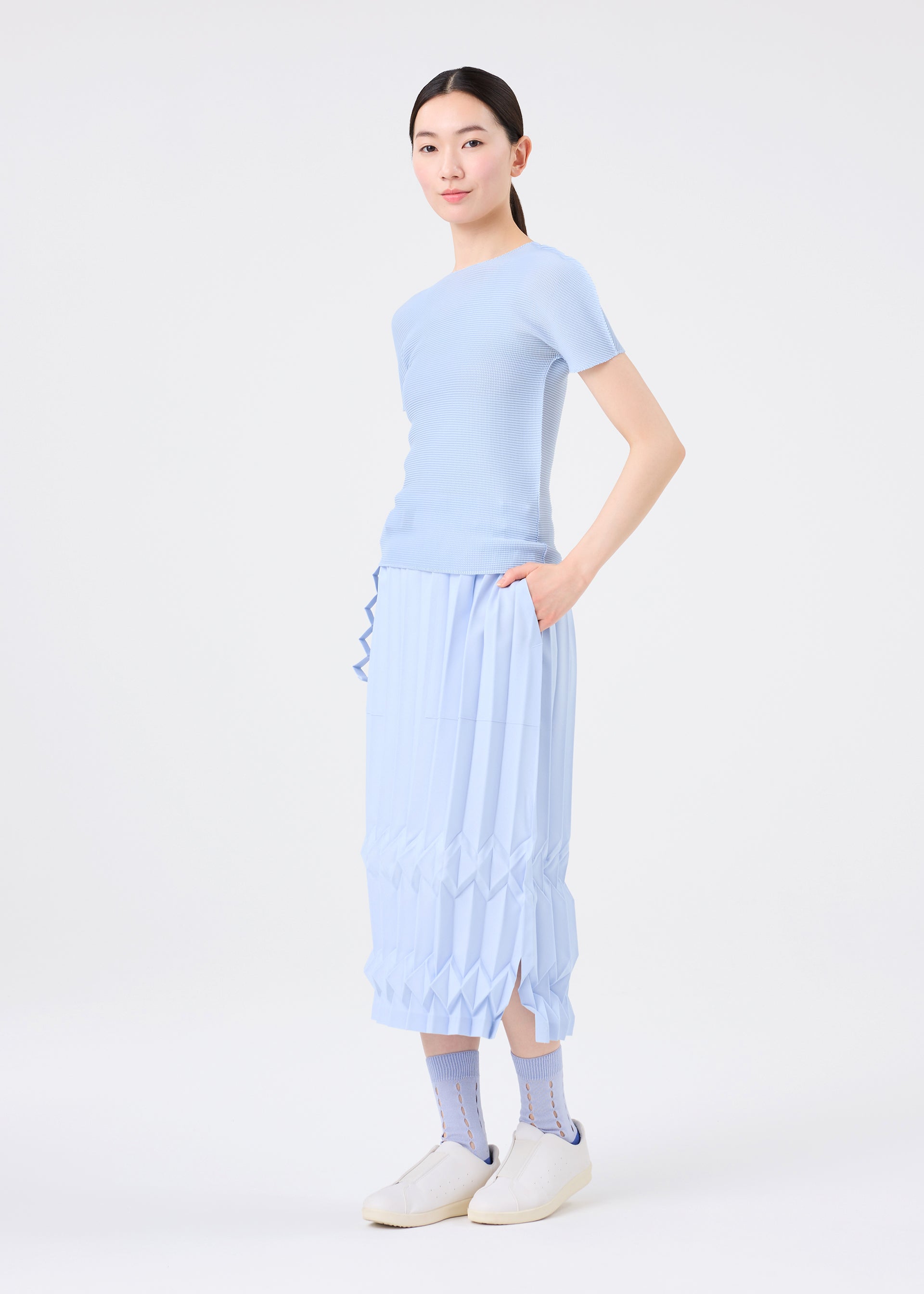Zoomed view A woman wears the TREE PLEATS skirt by me ISSEY MIYAKE with a light blue short-sleeve top, patterned blue socks, and white slip-on shoes. She stands with one hand in her pocket against a plain white background.