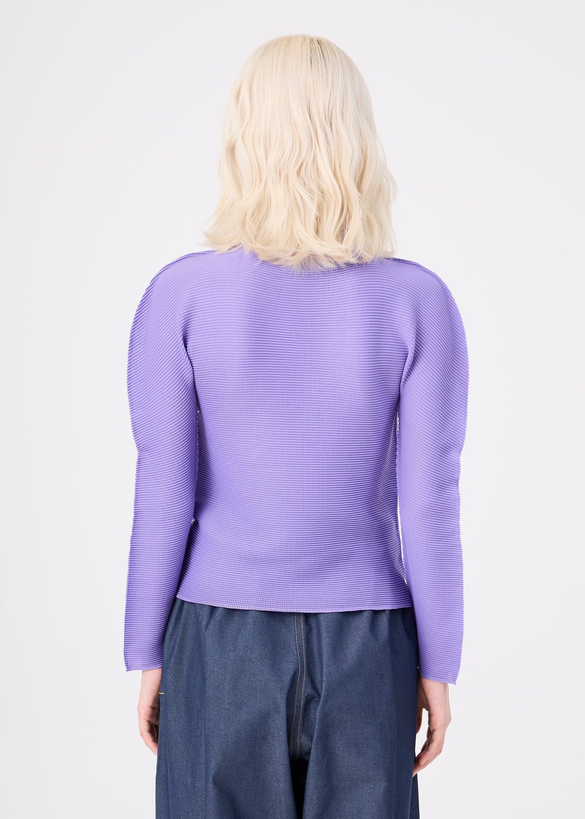 Zoomed view A person with shoulder-length blonde hair is seen from behind, wearing the me ISSEY MIYAKE STRETCH PLEATS 3 long-sleeved, high-neck top paired with high-waisted blue jeans against a plain white background.