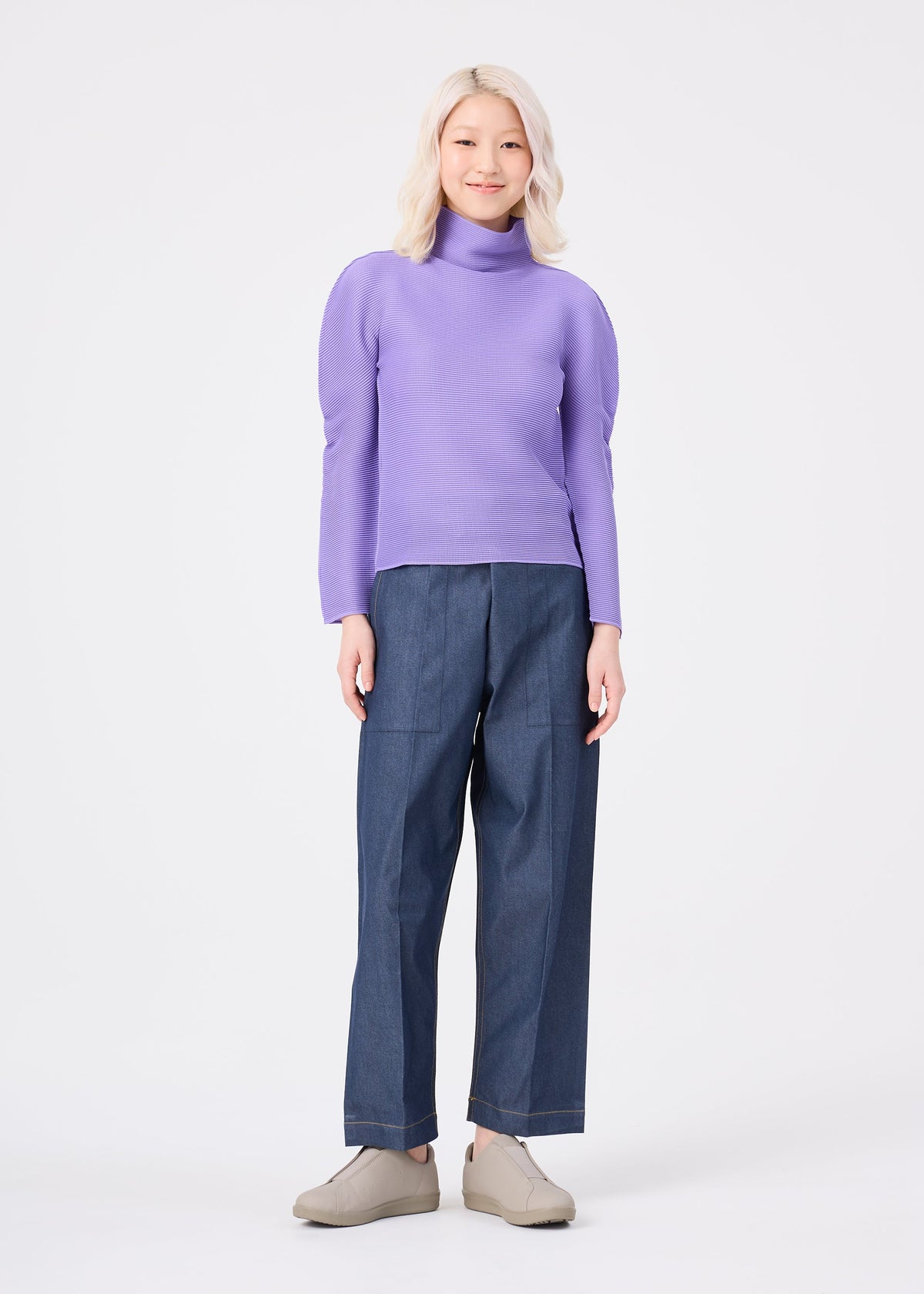 A person with light blonde hair is smiling against a plain white background while wearing the me ISSEY MIYAKE STRETCH PLEATS 3 long-sleeved lavender turtleneck, paired with wide-leg denim pants and beige shoes. view 7 of 7