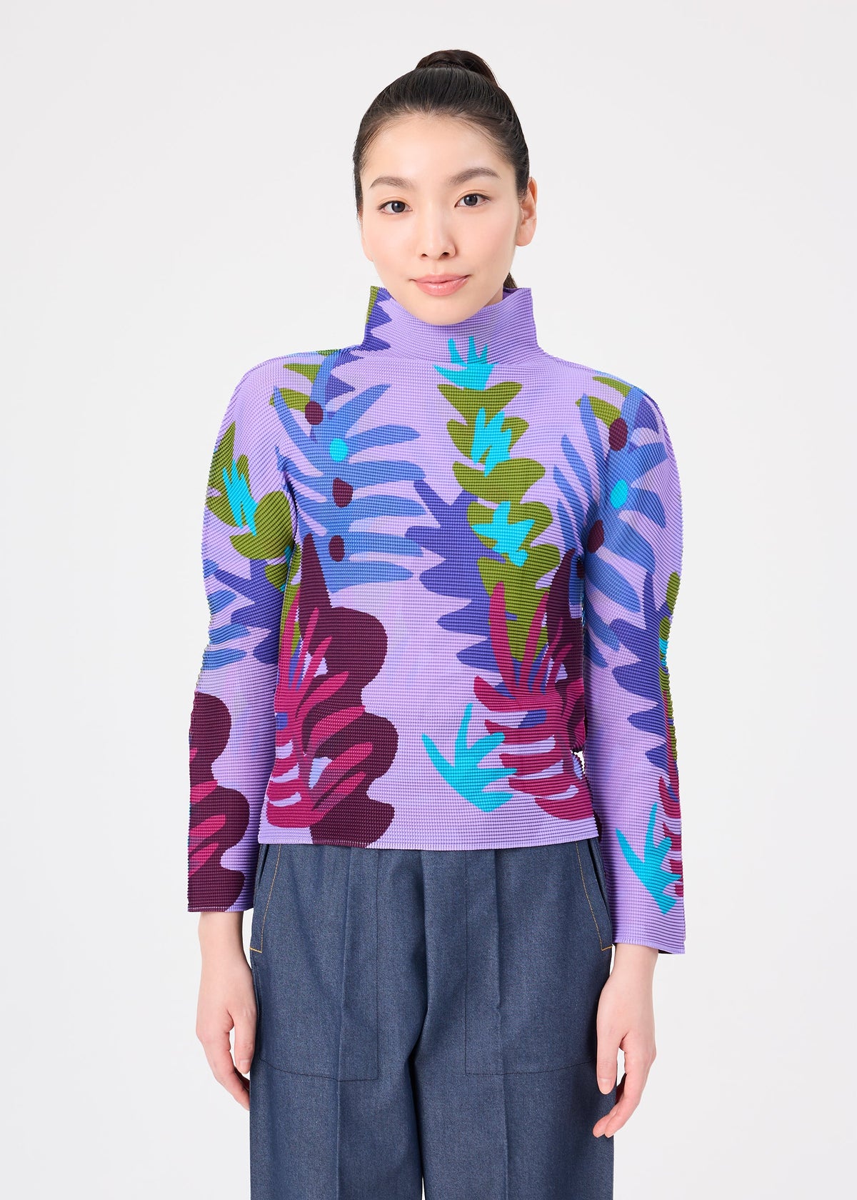 Zoomed view A person stands facing forward against a plain white background, wearing the “JUNGLE” high-neck long-sleeve top by me ISSEY MIYAKE, featuring bold and colorful leaf patterns, paired with loose-fitting blue pants.