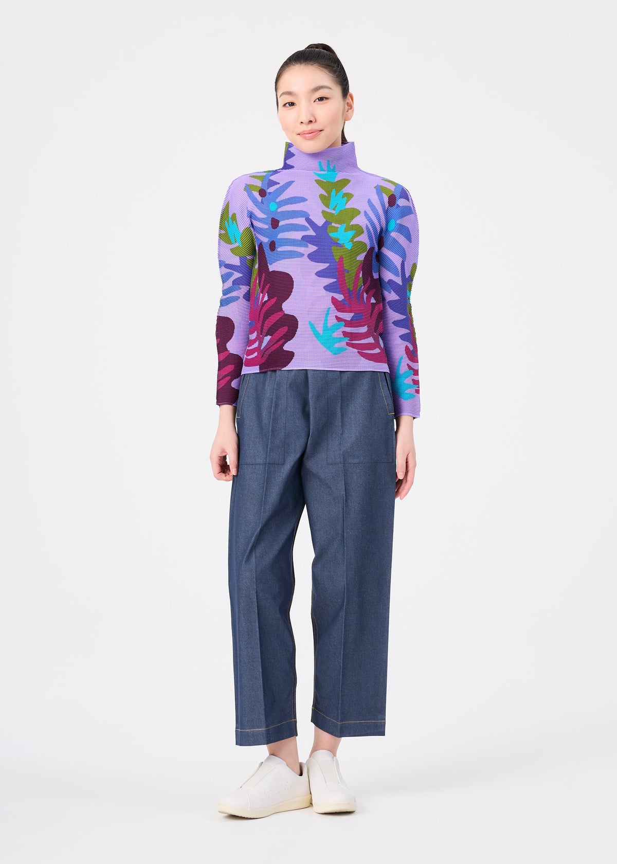 A person stands facing forward against a plain white background, wearing the me ISSEY MIYAKE JUNGLE high-neck long-sleeve sweater featuring a vibrant leaf pattern in purple, blue, and green tones, paired with loose blue pants and white shoes. view 6 of 6