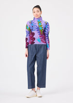 A person stands facing forward against a plain white background, wearing the me ISSEY MIYAKE JUNGLE high-neck long-sleeve sweater featuring a vibrant leaf pattern in purple, blue, and green tones, paired with loose blue pants and white shoes.