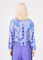 A person with wavy, shoulder-length blonde hair is seen from behind, wearing the PLANT high-neck long-sleeve top by me ISSEY MIYAKE, which features abstract blue and turquoise plant patterns, paired with pleated purple pants. The background is plain white.