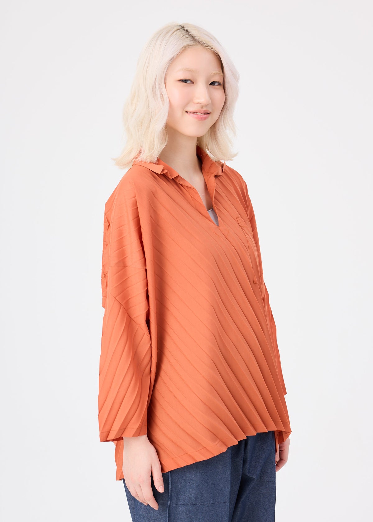 A woman with light blonde hair stands smiling against a plain white background, wearing the ANGLE PLEATS SHIRT by me ISSEY MIYAKE in orange and blue pants. view 6 of 8