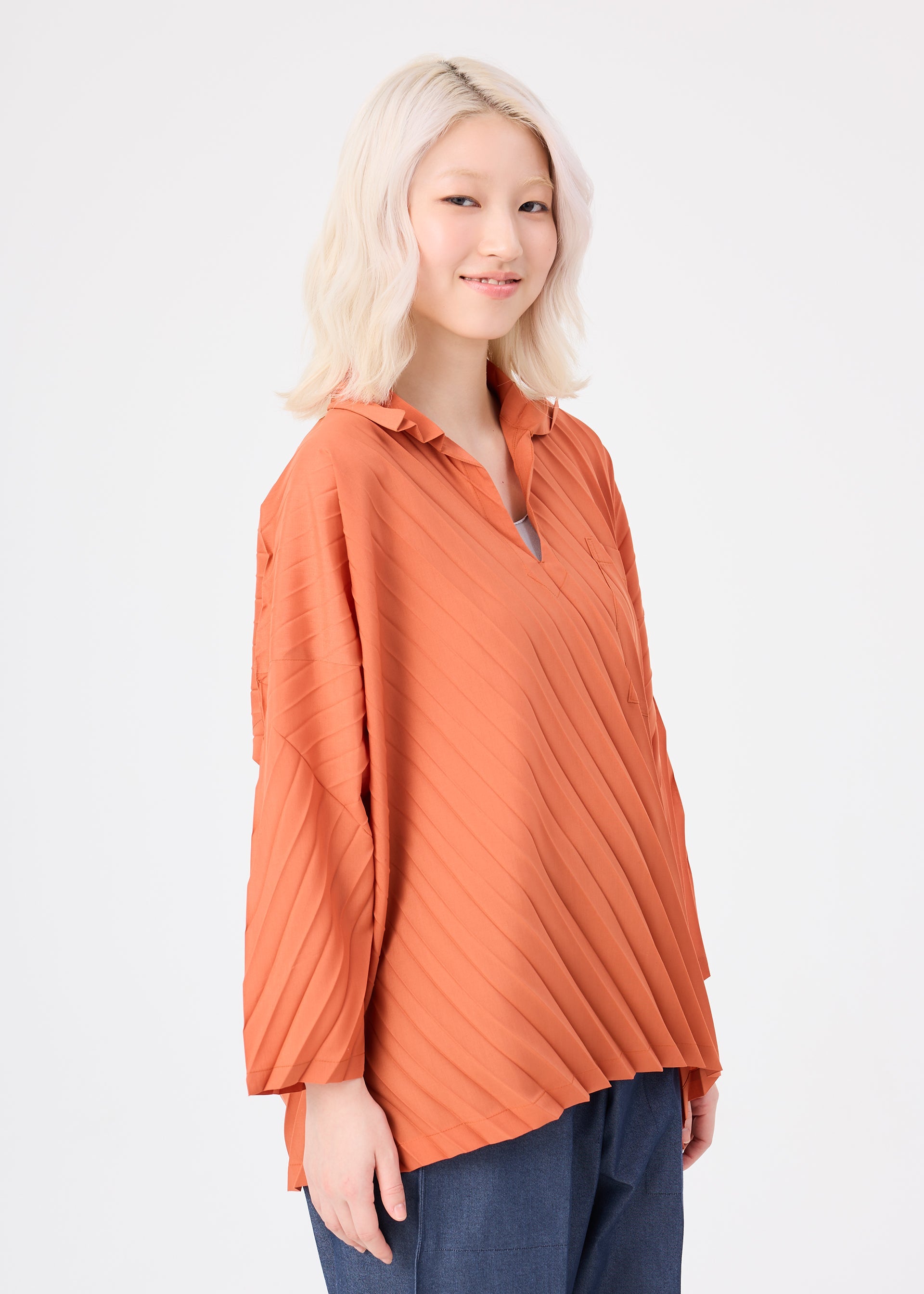 Zoomed view A woman with light blonde hair stands smiling against a plain white background, wearing the ANGLE PLEATS SHIRT by me ISSEY MIYAKE in orange and blue pants.