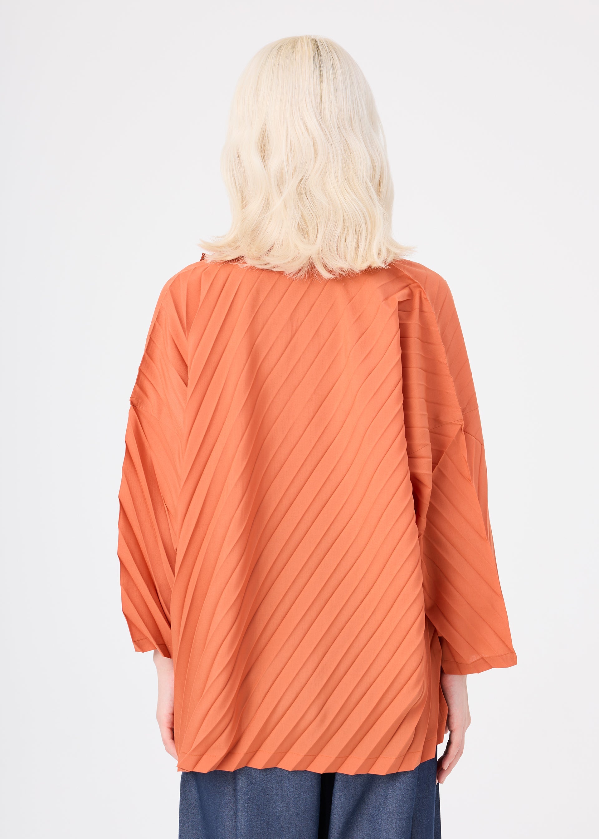 Zoomed view A person with shoulder-length, wavy platinum blonde hair is seen from behind wearing the ANGLE PLEATS SHIRT by me ISSEY MIYAKE—an oversized, orange, textured three-quarter-sleeve shirt with diagonal pleats—paired with loose-fitting blue pants against a plain white background.