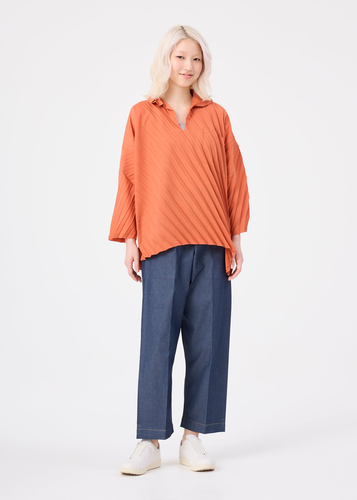 A person with light blonde hair wears the me ISSEY MIYAKE ANGLE PLEATS SHIRT in orange, styled with blue wide-leg pants and white shoes, standing against a plain white background. view 8 of 8
