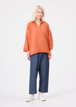 A person with light blonde hair wears the me ISSEY MIYAKE ANGLE PLEATS SHIRT in orange, styled with blue wide-leg pants and white shoes, standing against a plain white background.