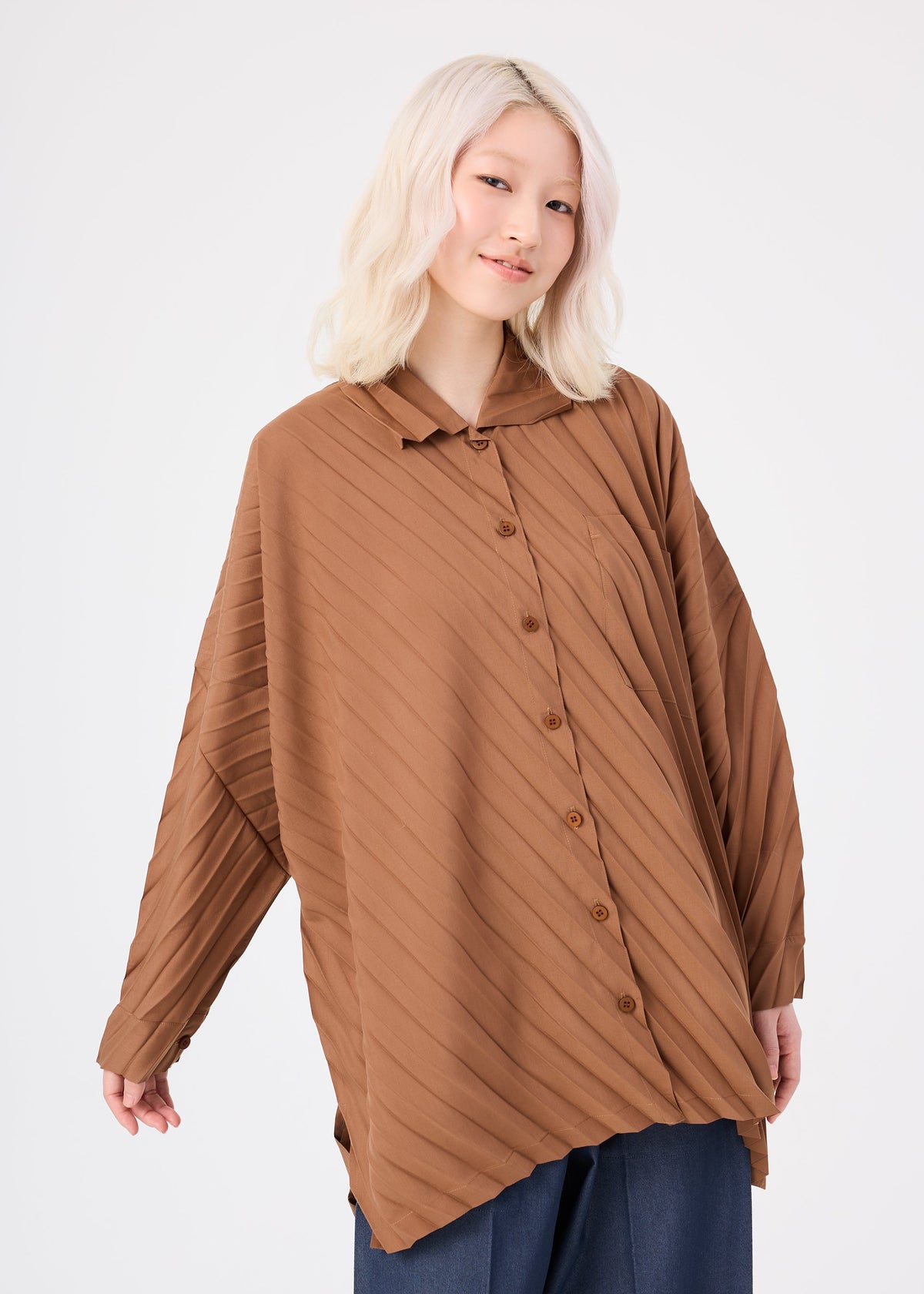 A person with light blonde hair wears the me ISSEY MIYAKE ANGLE PLEATS SHIRT, a loose-fitting, brown, long-sleeved top with diagonal stripes, paired with dark pants. They stand against a plain white background and smile gently at the camera. view 6 of 8