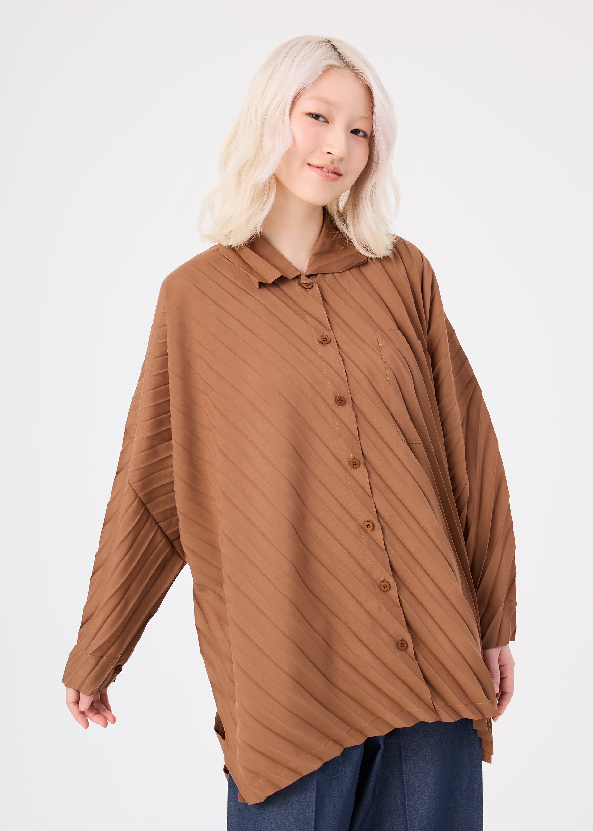Zoomed view A person with light blonde hair wears the me ISSEY MIYAKE ANGLE PLEATS SHIRT, a loose-fitting, brown, long-sleeved top with diagonal stripes, paired with dark pants. They stand against a plain white background and smile gently at the camera.