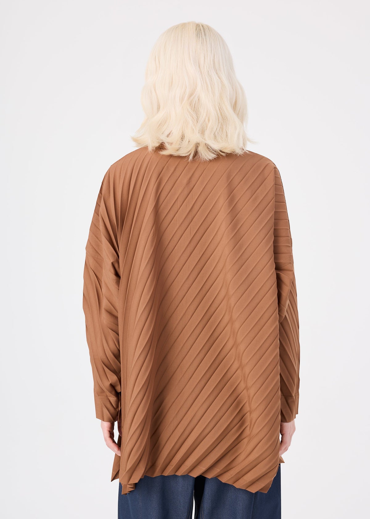 A person with shoulder-length blonde hair is shown from the back, wearing the ANGLE PLEATS SHIRT by me ISSEY MIYAKE—a loose, brown, pleated, long-sleeved shirt with diagonal stripes. They are also wearing dark pants. The background is plain white. view 7 of 8