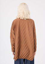 A person with shoulder-length blonde hair is shown from the back, wearing the ANGLE PLEATS SHIRT by me ISSEY MIYAKE—a loose, brown, pleated, long-sleeved shirt with diagonal stripes. They are also wearing dark pants. The background is plain white.