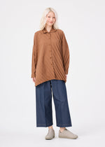 A person with light hair wears the me ISSEY MIYAKE ANGLE PLEATS SHIRT, a loose and versatile long-sleeved brown button-up, paired with wide blue jeans and light shoes, standing against a plain white background.