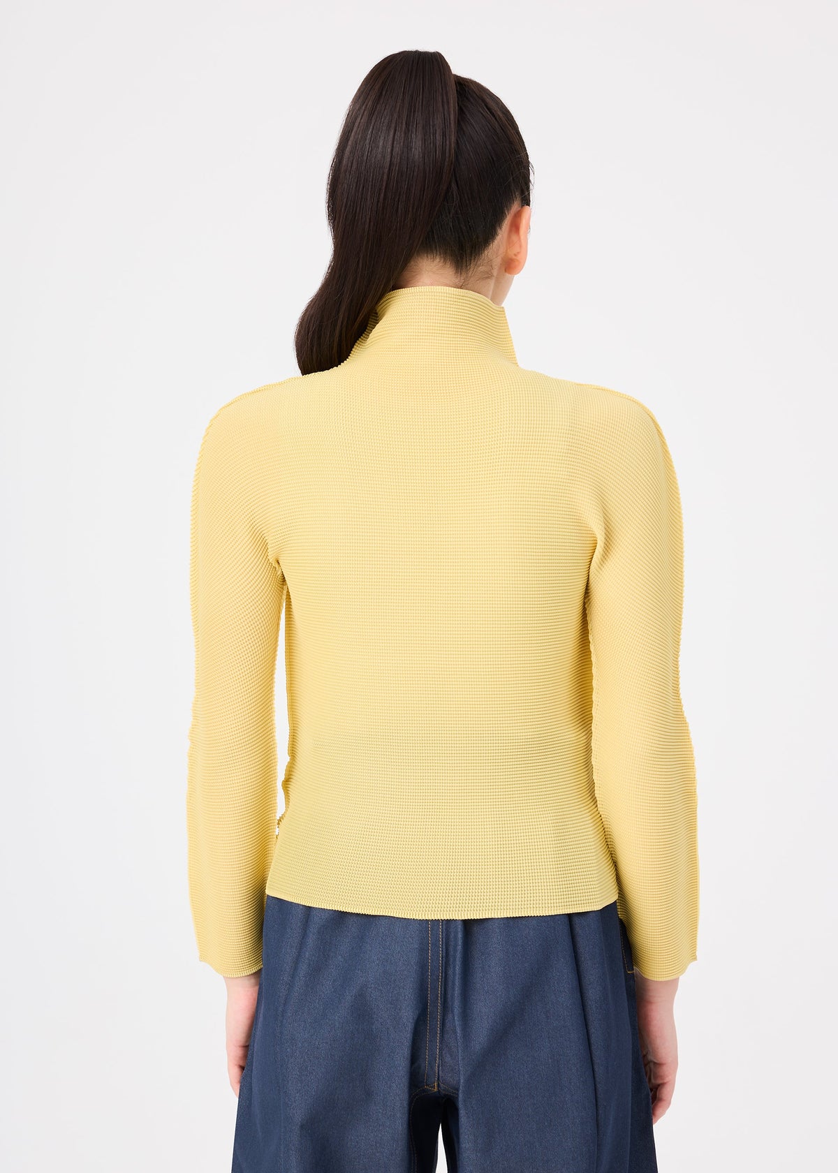 Zoomed view A person with long, dark hair in a ponytail wears the yellow STRETCH PLEATS 4 top by me ISSEY MIYAKE and blue jeans, standing with their back to the camera against a plain white background.