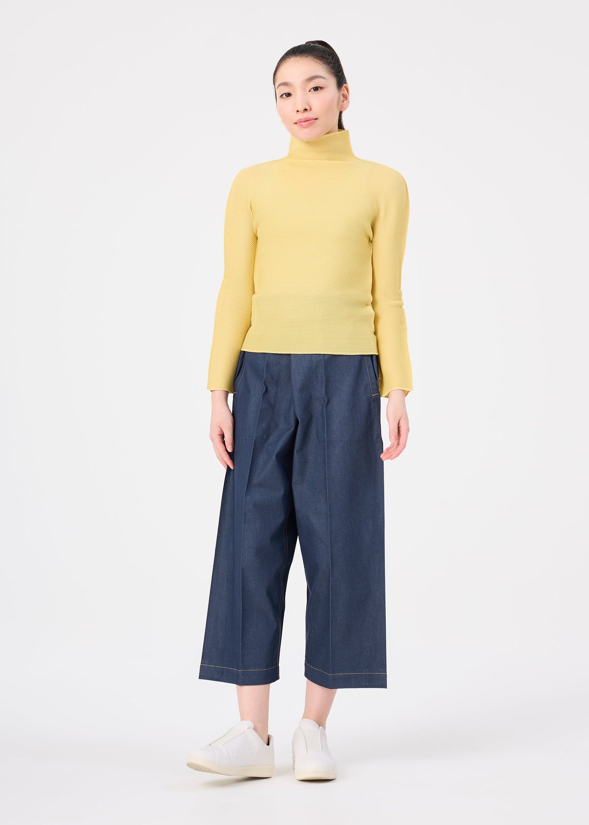 A woman stands against a white background wearing the STRETCH PLEATS 4 top by me ISSEY MIYAKE, paired with wide-leg blue denim pants and white sneakers. Her hair is tied back, arms relaxed at her sides. view 7 of 7