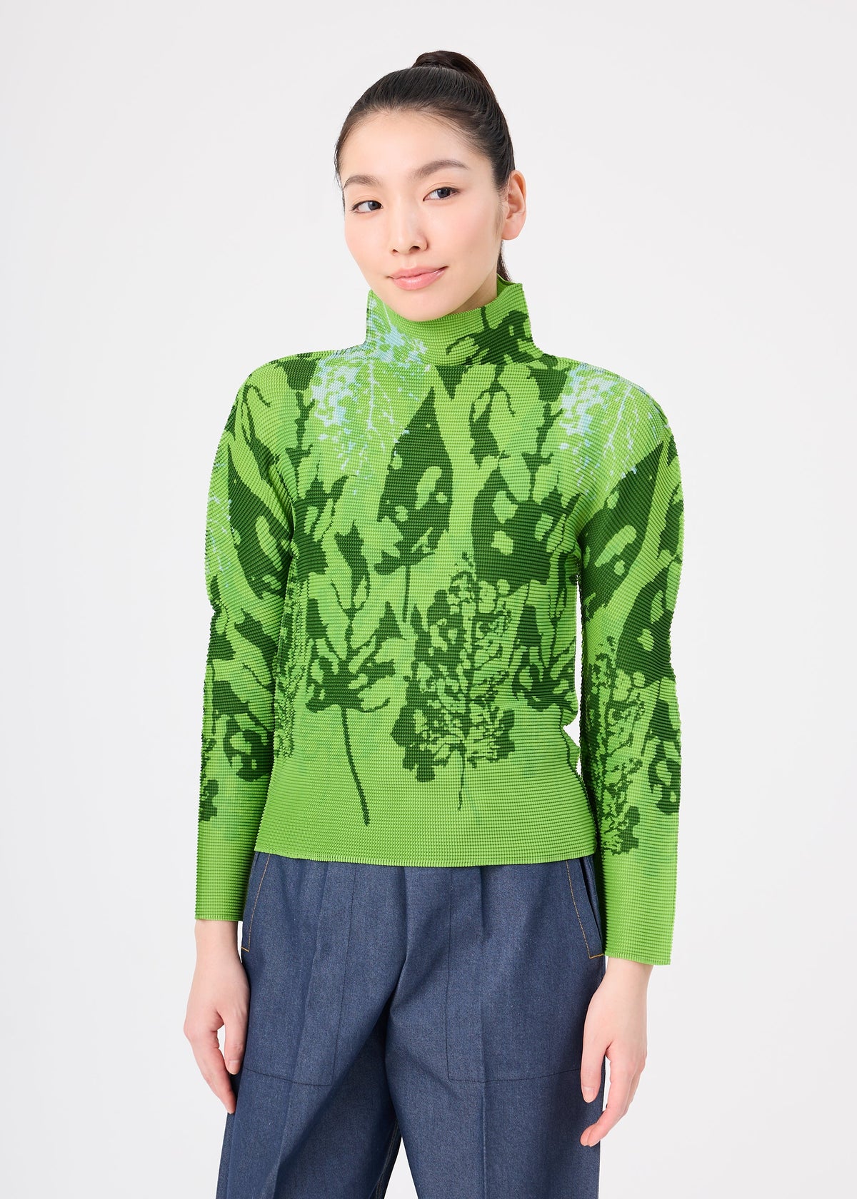 A woman with long dark hair in a high ponytail wears the LEAF shirt by me ISSEY MIYAKE, paired with a green high-neck top and blue high-waisted pants against a plain white background. view 5 of 7